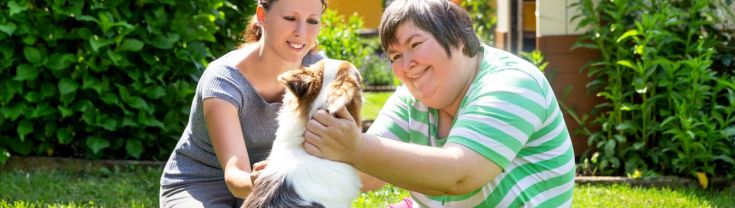 Woman with intellectual disability sitting on grass with carer patting dog