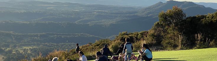 People sitting on grass overlooking the bushland below with mountains in the background