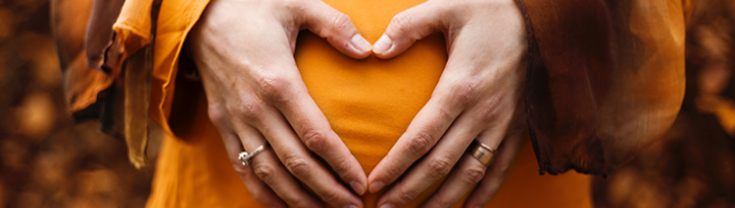 Close up of pregnant belly in orange shirt, with hands held together to make a heart shape in front