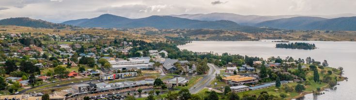 Aerial shot over Jindabyne buildings with water in foreground, mountains behind
