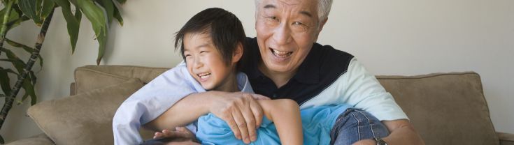 Image of three smiling people - an older Asian man with greying hair smiling and two younger Asian children. The grandfather's arm is around the child on his right and the second child is laying across his lap laughing. They are sitting on a brown suede couch and the is a tall green plant on their right. 