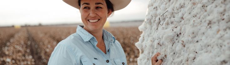 A woman is standing in a paddock up against a round bale of hay. She is wearing a light blue shirt, and cream, wide brimmed hat and is smiling.
