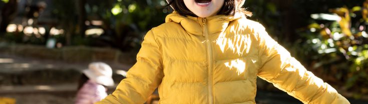 A young girl of 4 or 5 is standing in a park on a sunny day wearing a yellow hat and yellow puffer jacket. She is smiling.