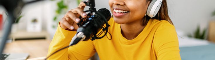A young woman in a yellow long sleeve top wears white headphones and speaks into a suspended black microphone