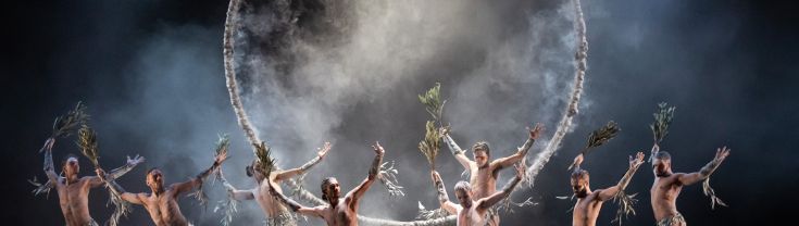 First nations dancers on stage filled with smoke, holding and covered in Australian native bush and white markings. 