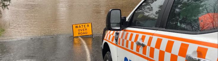 A State Emergency Services car is stopped in front of a sign saying "water over road", and a flooded road.