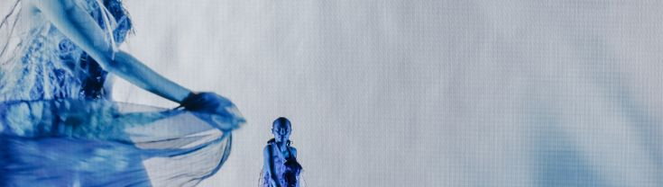 Model twirling her dress on a stage of blue lighting with a projection of her behind her on a screen .