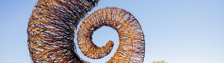 A snail shell like spiral sculpture, made out of steel, freestanding in an outdoor landscape against a blue sky.