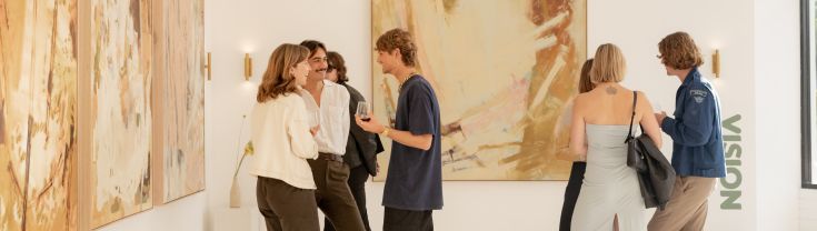 A small crowd of young people inside a gallery holding drinks and chatting. 