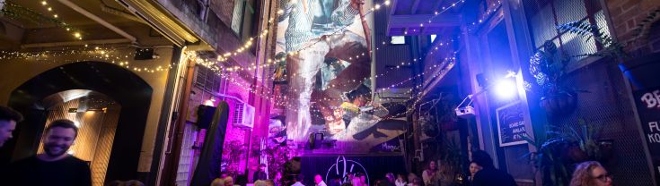 A bustling laneway scene with large art work on the walls, lots of people and fairy lights hanging above everyone. 