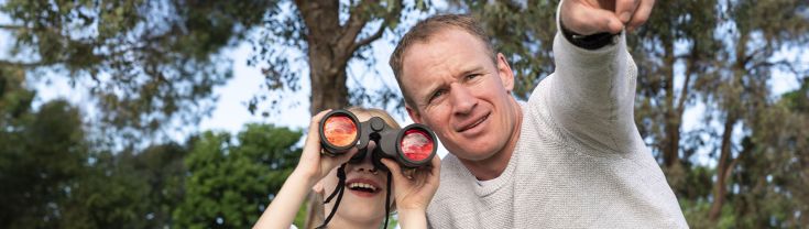 A man wearing a grey long sleeve jumper points to something behind the camera. A young girl in a blue shirt and shorts looks through binoculars she is holding