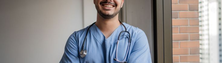 A male nurse stands in his office, smiling with arms folded.