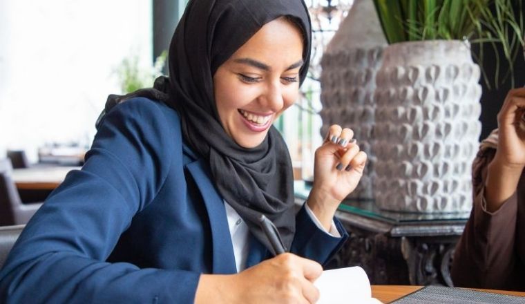 A smiling woman wearing a business suit and hijab writing on a notepad