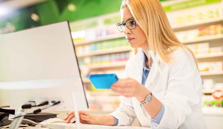 A blonde pharmacist wearing a white coat, holding a medication packet and looking at a computer.