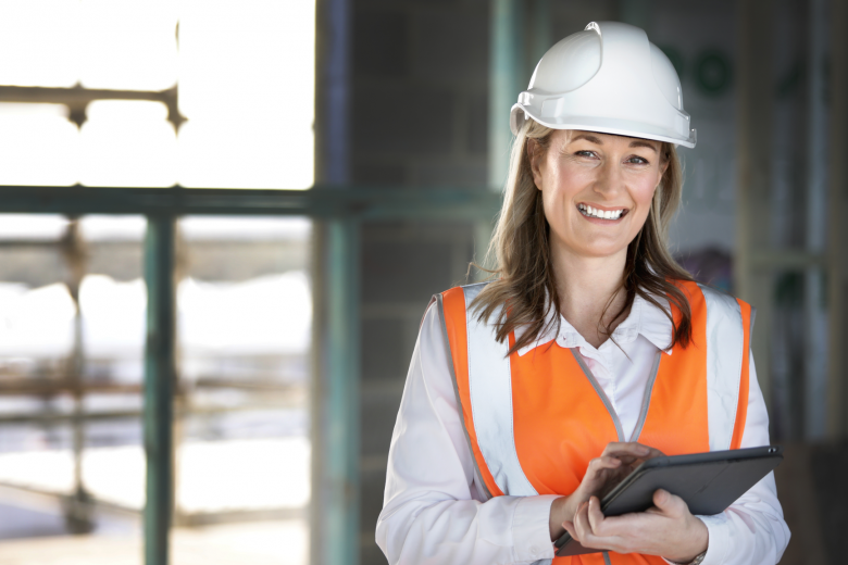 A woman is standing in a hi-vis vest and a hard hat with a clip board in her arms. 