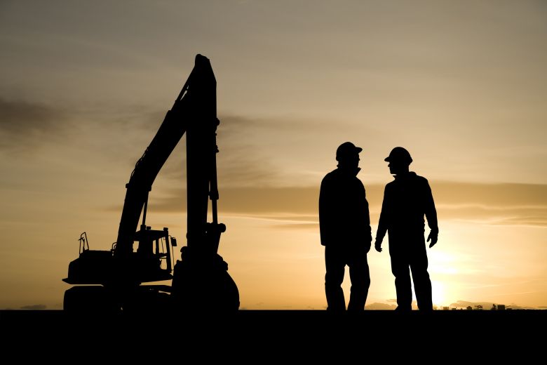 Silhouette of two workers wearing hard hats near a large digger on a mine site.