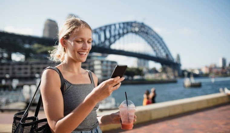 A female holding a mobile phone standing outside with the Sydney Harbour Bridge in the background. 