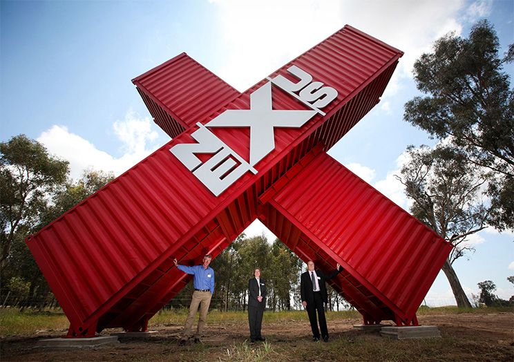 3 men standing under the red sign made out of crossed containers outside Nexus Industrial Job Precinct