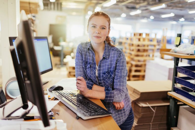 Young woman in blue shirt seated at desktop monitor with open plan office in background.