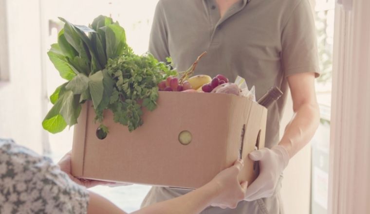 A man handing a box of fruit and vegetables to a lady at her front door.