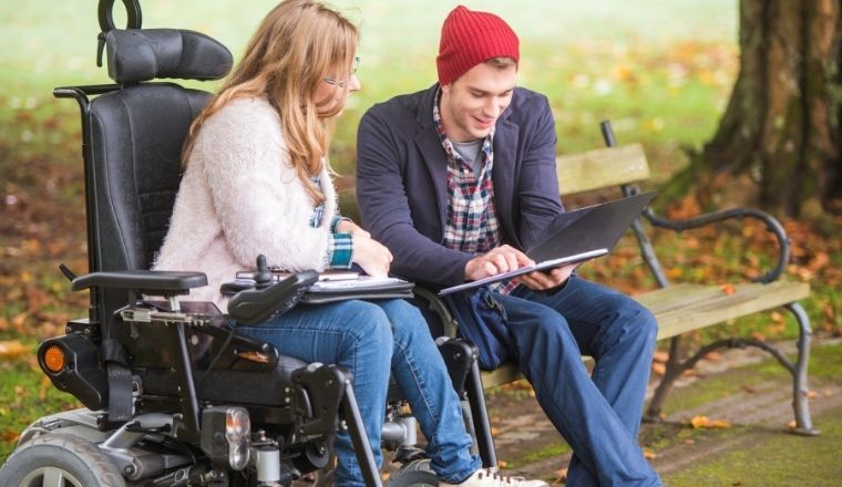 Woman in a wheelchair talking to a man who is seated with a laptop computer on his lap.