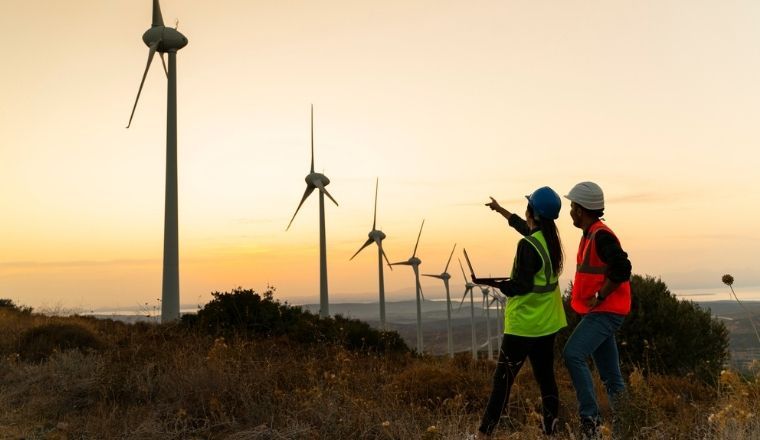 A man and a woman in PPE standing in front of wind turbines at sunset.