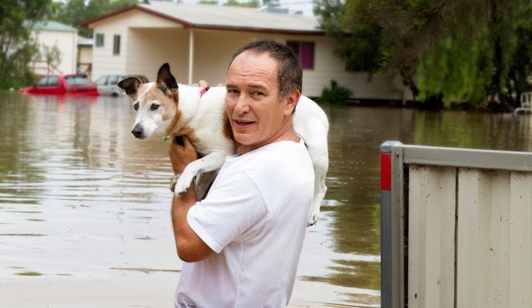 A man walking through flood waters while holding his dog.
