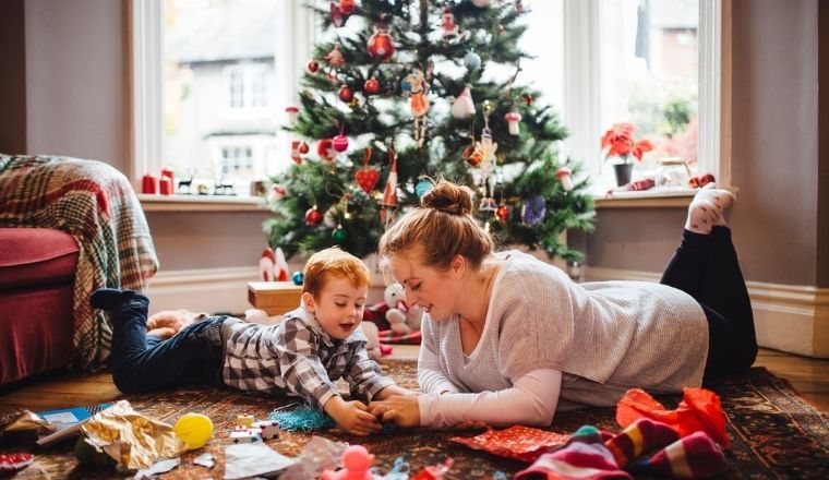 A mother and young boy lying on the floor in front of a Christmas tree, playing with newly opened toys together.