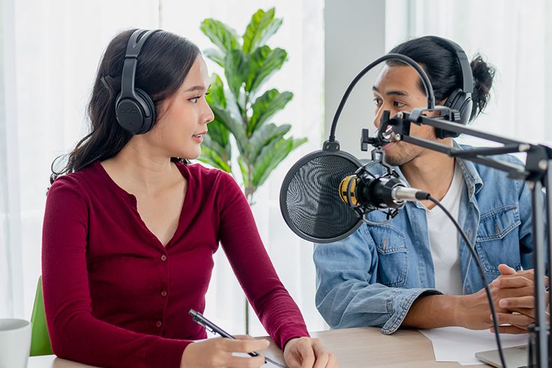 A young woman wearing headphones talking to a young man also wearing headphones behind a microphone which is covered by a pop filter