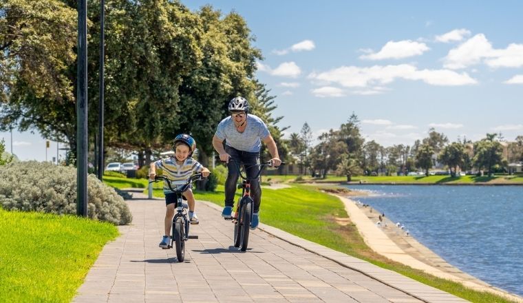 A young boy and father both riding bicycles on a cycling track next to a lake.