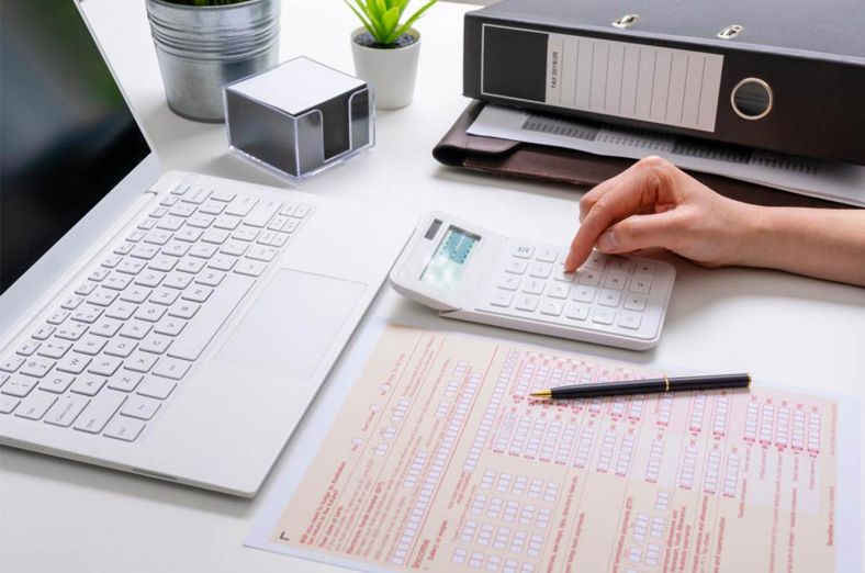Tax form, folder and laptop on a desk with a hand using a calculator