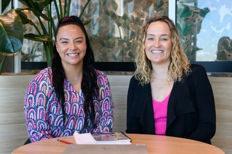 Two women sitting in a breakout area smiling at the camera.
