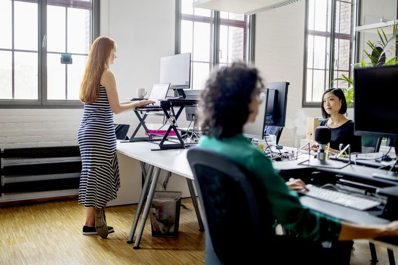 Woman standing and working a ergonomic desk in modern office with colleagues working around. 