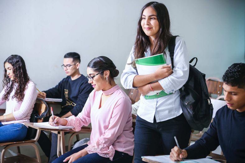 Young adult students sit at classroom desks with a woman with a back and holding a notebook standing and smiling