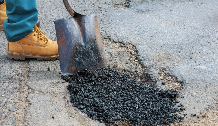 A pothole in the road, being repaired by a workman with a shovel.