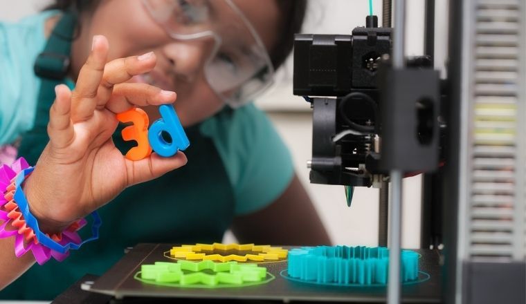 A young female student wearing multi-colored 3D printed shapes as jewelry, next to a 3D printer with designs on the heated print bed, and holding 3d letters in her hand.