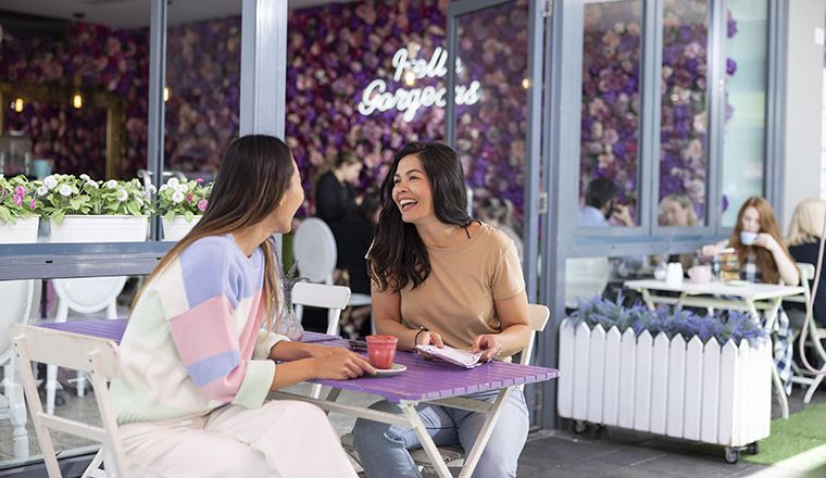 The image shows two women sitting at the Social Hideout Cafe in Parramatta enjoying drinks. The cafe features very bright purple and pink flowers with a neon sign that says 'hello gorgeous' in the background of the image