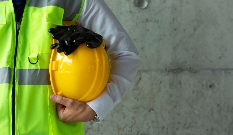 A close-up photo of a man wearing a fluro vest and holding workplace Personal Protection Equipment, consisting of a yellow helmet, and protective gloves.