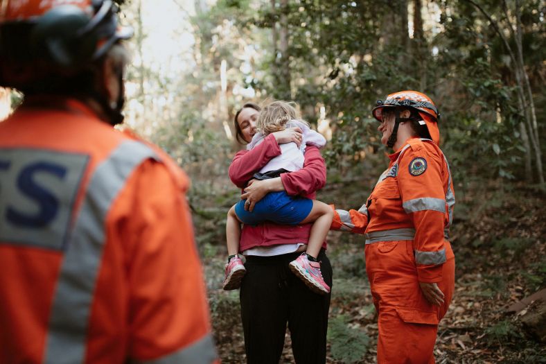 Adult holding child in centre while two volunteers in orange SES gear and helmets look on.