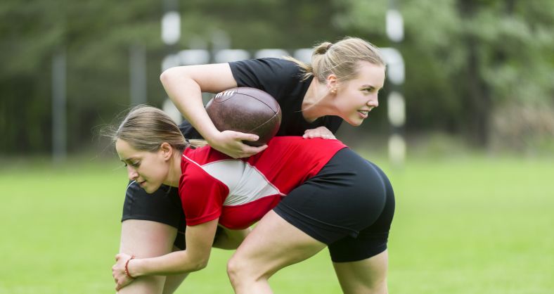 Two women rugby players practising a tackle drill with a rugby ball.