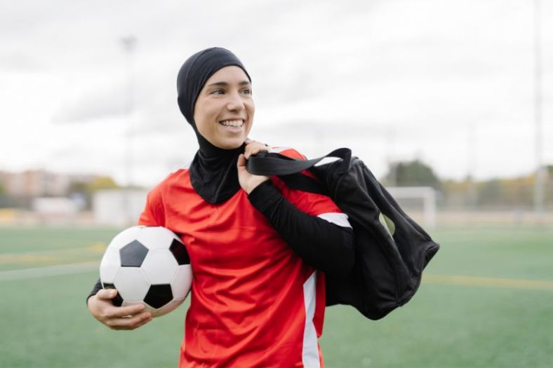 A muslim woman in a head scarf and athletic gear holding a soccer ball