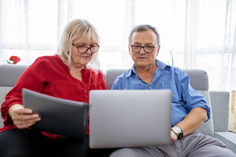 Male and female couple on couch with a laptop and paperwork doing their strata annual reporting.