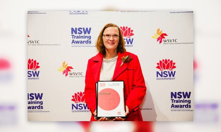 Smiling woman in bright red jacket stands holding a framed certificate, NSW Government and NSW Trauing Awards on banner backdrop
