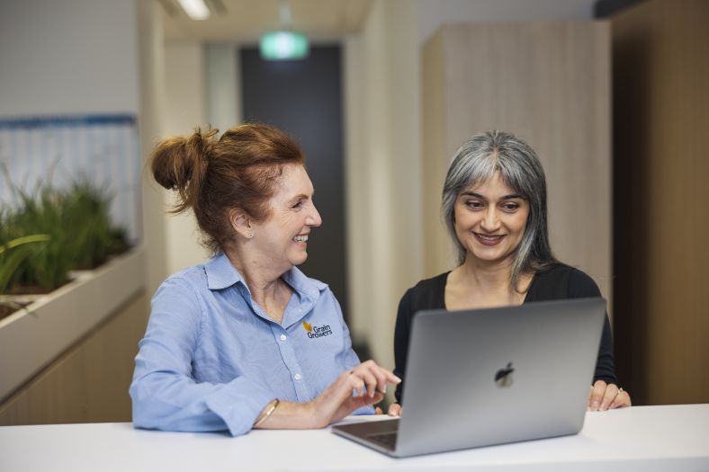Woman with red hair wearing a blue shirt standing next to a woman with grey hair wearing a black shirt. Both are smiling while working on a grey laptop.