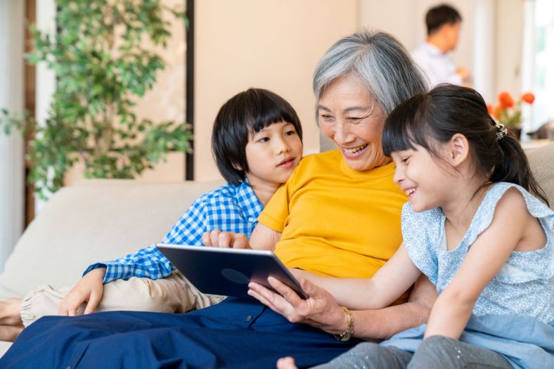 Image of Asian grandmother with her grandson and granddaughter on either side of her. Her grandson is looking her her and her granddaughter is smiling, looking at the iPad her grandmother is also looking and smiling at. They are sitting on a cream-coloured couch. A green tree is to their right and another man is seen blurred in the background. 