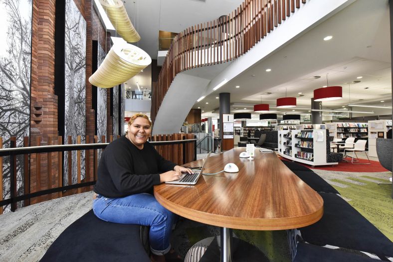 A young aboriginal woman in university library smiling a the camera with a laptop ion front of her.