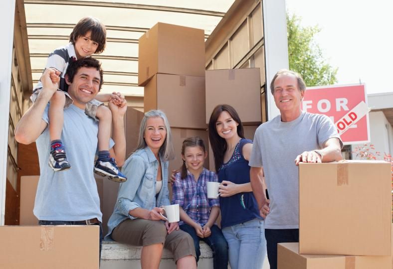 A multi-generational family sitting on the back of a truck with moving boxes and a sold sign in the background