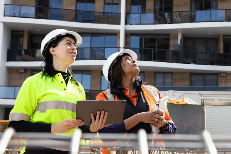 Two inspectors wearing white hard hats, one wearing a yellow vest and holding a clipboard, and the other wearing an orange vest, looking up at a building.