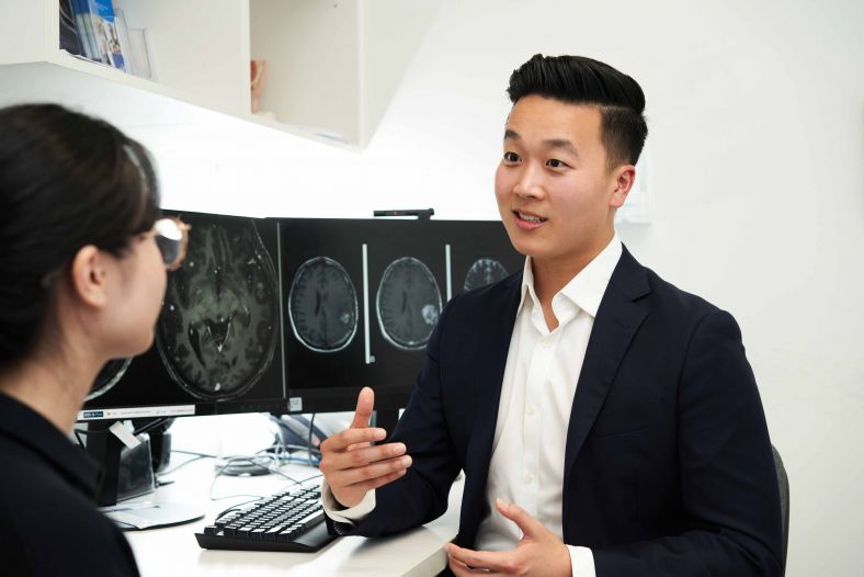 Dr Kevin Jang speaking with a colleague in front if dual computer monitors showing cross-sectional black and white images of human brain