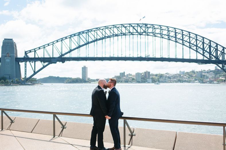 A couple kiss on the Sydney Opera House forecourt with the harbour bridge in the backgroundr 